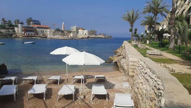 A beachside area with white lounge chairs and umbrellas, a stone wall, and a view of the sea and buildings in the background. - 24/7 electricity Seaview studio with beach access (Jabal Lubnan)