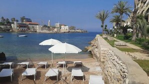 Vista de la playa con sillas de descanso y sombrillas, un muro de piedra y un mar tranquilo con barcos.