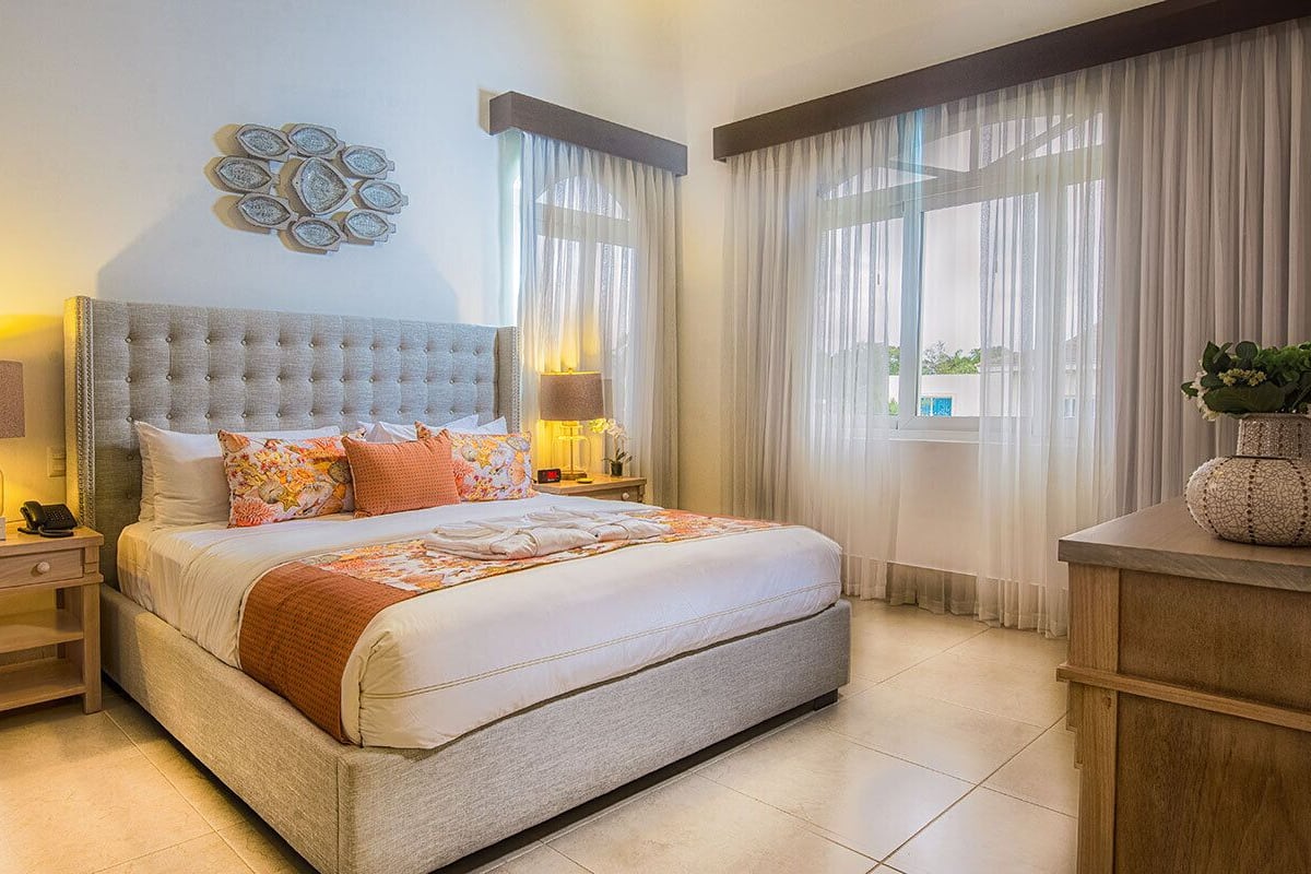 A bedroom with a large bed, two bedside tables with lamps, a decorative wall piece, a window with sheer curtains, and a small plant on a side table.