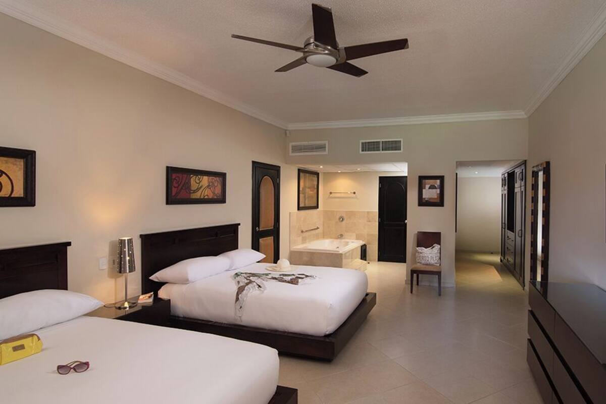 A hotel room with two beds, a ceiling fan, a TV, and a bathroom with a bathtub and shower.