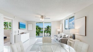 A spacious living room with a glass coffee table, white furniture, a ceiling fan, a TV on the wall, and a view of the outdoors. - Boca Raton - Across from Beach - Balcony • Pool • Parking • 2BR (Boca Raton)