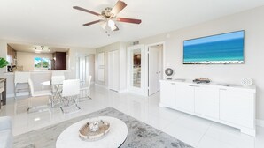 A modern living room with a marble coffee table, white furniture, a ceiling fan, and a large TV displaying a beach scene. - Boca Raton - Across from Beach - Balcony • Pool • Parking • 2BR (Boca Raton)