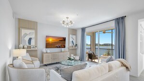 Una sala de estar moderna con sofá, sillones, una mesa de centro y televisor. Hay un balcón con vista al agua.