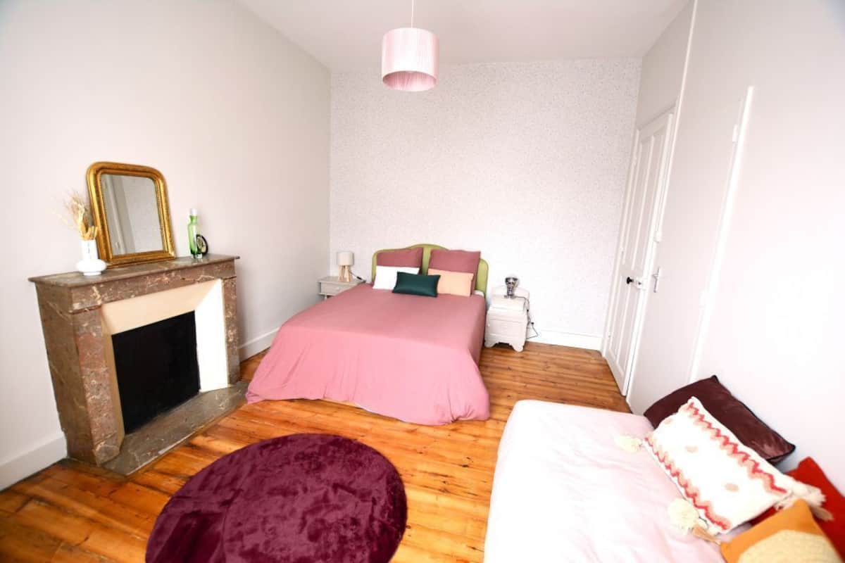 A bedroom with a wooden floor, a bed with a pink bedspread, a fireplace, and a mirror on the wall.