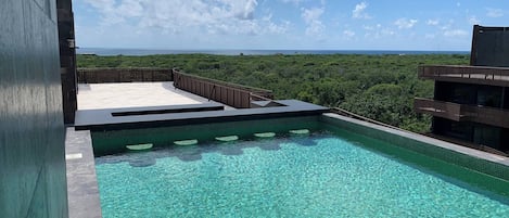 A rooftop pool with clear blue water, surrounded by lush greenery and a view of the ocean.
