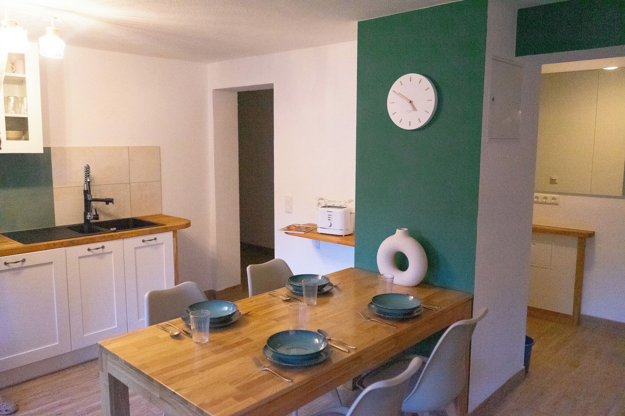 Cocina compacta con una mesa de madera preparada para comer, un reloj blanco en la pared y una pared de acento verde.