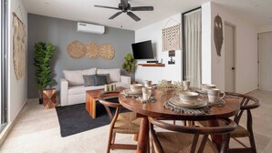 A modern living room with a round wooden dining table set for four, a grey sofa, a ceiling fan, and a TV mounted on the wall. - Beautiful and Comfortable, 1 Bedroom! The Best in Zama! (Tulum)