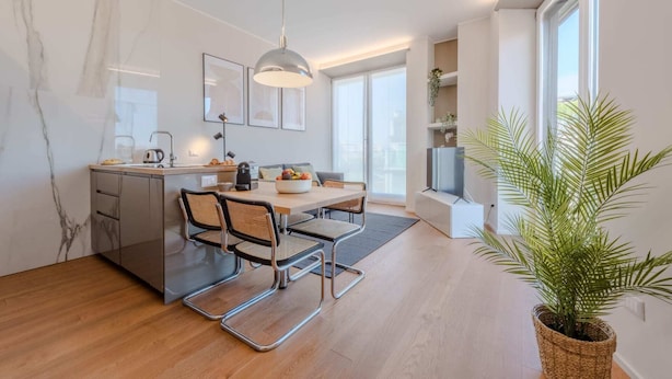 A modern kitchen with a dining area, wooden flooring, and a large window providing natural light.