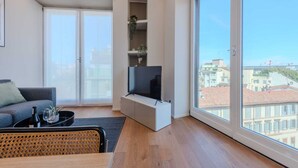A modern living room with a sofa, a coffee table, a TV, and a large window offering a city view.