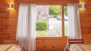 Una cabaña de madera con techo alto, un comedor con mesas y sillas de madera, y un ventanal con cortinas.