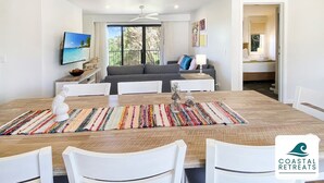 A modern dining area with a wooden table, white chairs, and a colorful rug. A gray sofa and a television are visible in the background. - 🏝 Luxury Poolside Retreat – Ramada Resort (Cowes)