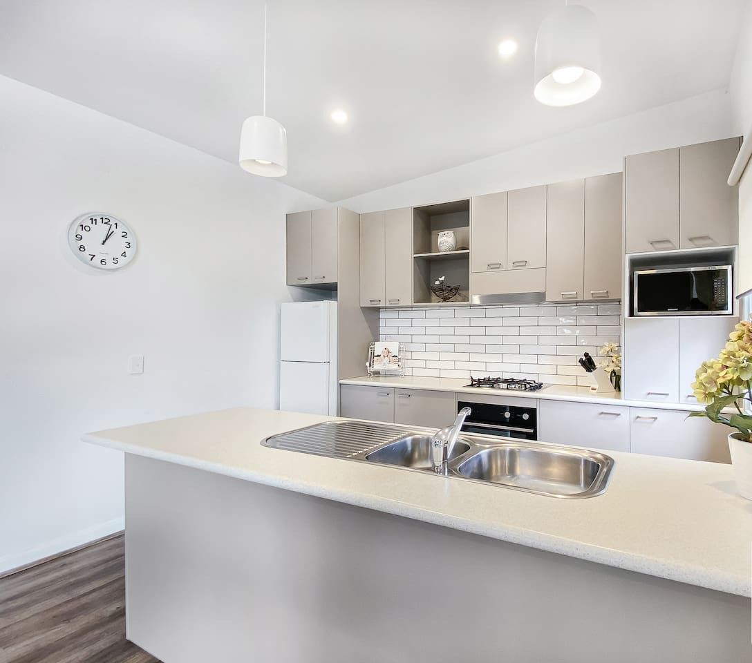 Une cuisine moderne avec un îlot central, des meubles blancs, un four à micro-ondes et un évier. Il y a une horloge au mur et une plante sur le comptoir.