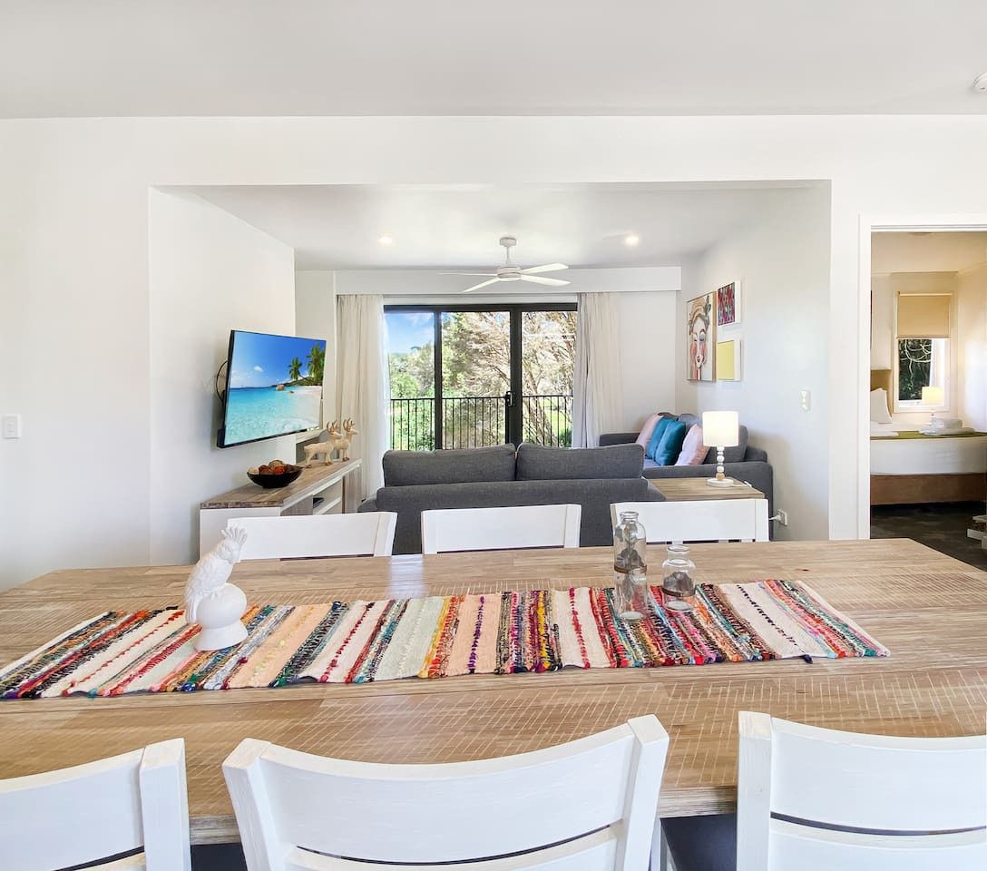Un espace repas moderne avec une table en bois, des chaises blanches et un tapis coloré. Un canapé gris et un téléviseur sont visibles à l’arrière-plan.