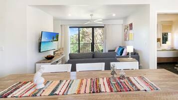 Un espace repas moderne avec une table en bois, des chaises blanches et un tapis coloré. Un canapé gris et un téléviseur sont visibles à l’arrière-plan.