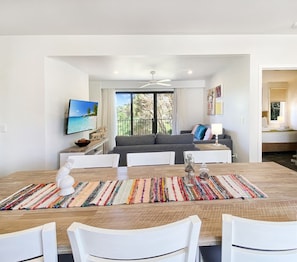 Un comedor moderno con una mesa de madera, sillas blancas y una alfombra colorida. Se ve un sofá gris y un televisor al fondo.