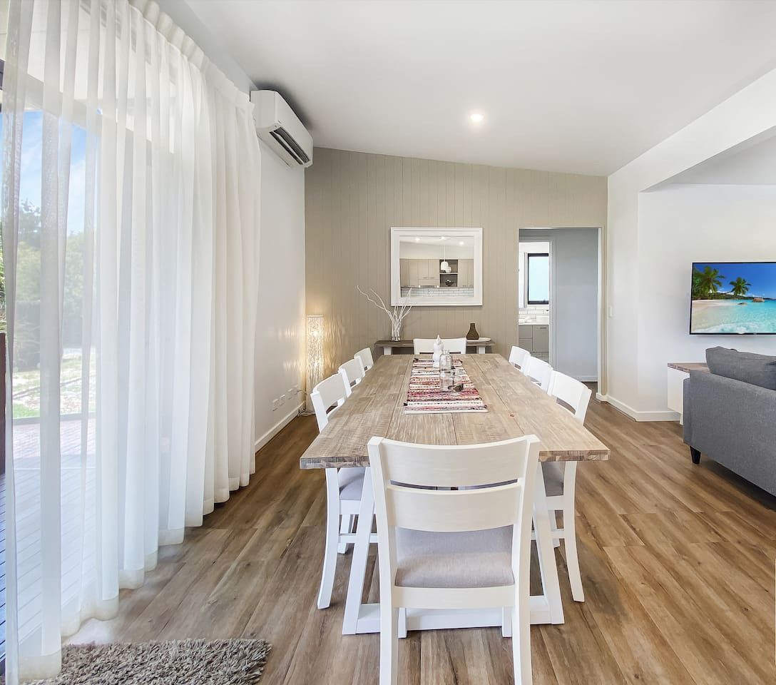 A modern living room with a wooden dining table, white chairs, a grey sofa, and a TV on the wall.