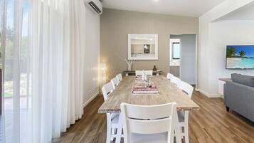 A modern living room with a wooden dining table, white chairs, a grey sofa, and a TV on the wall.