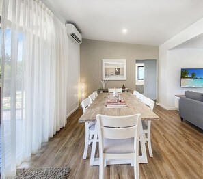 Un salon moderne avec une table à manger en bois, des chaises blanches, un canapé gris et un téléviseur fixé au mur.