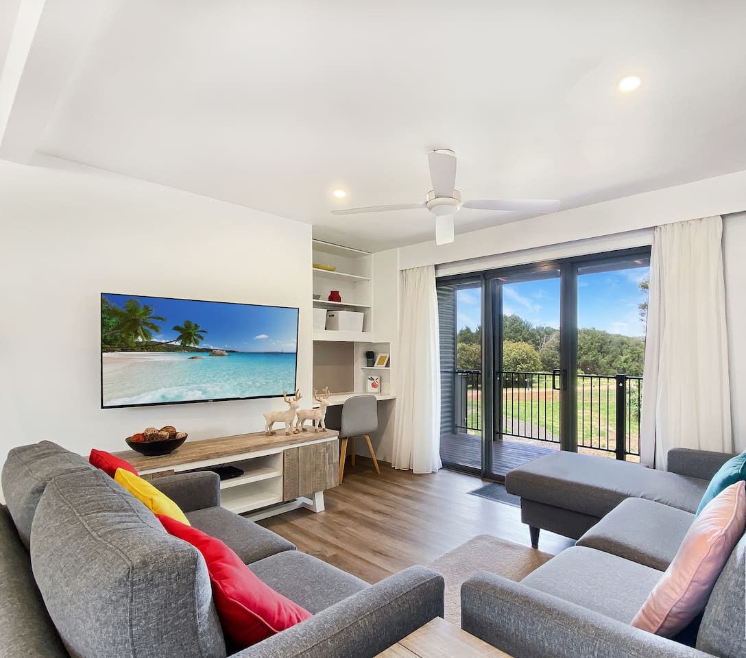 A modern living room with a flat-screen TV, grey sofas, a wooden coffee table, and a balcony with a view of the outdoors.