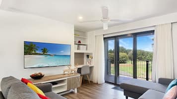A modern living room with a flat-screen TV, grey sofas, a wooden coffee table, and a balcony with a view of the outdoors.