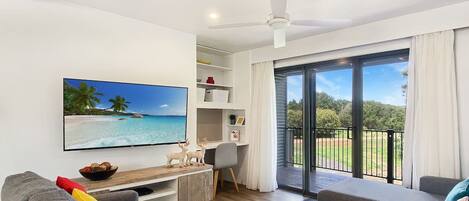 A modern living room with a flat-screen TV, grey sofas, a wooden coffee table, and a balcony with a view of the outdoors.