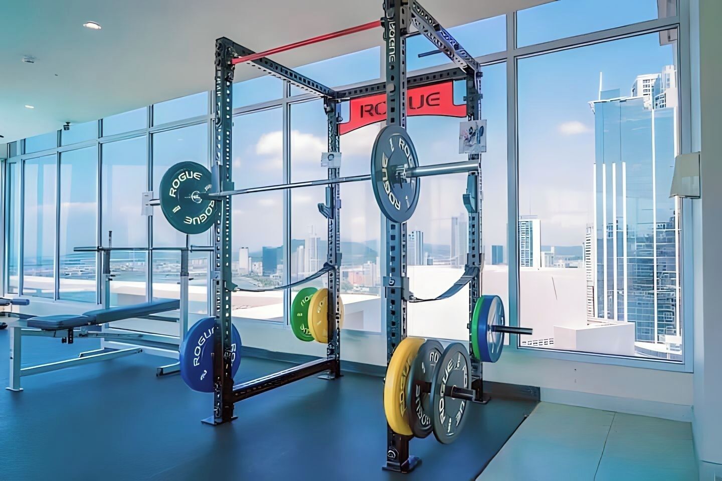 Un espace dédié à la musculation, équipé d’un support pour les squats et de divers disques de poids, de grandes fenêtres offrant une vue sur la ville, ainsi que d’un banc.