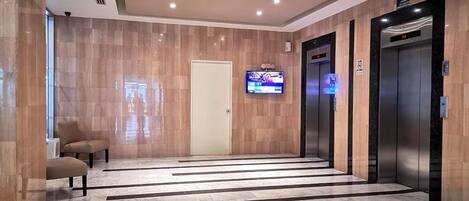 Un vestíbulo de ascensores moderno con suelo de mármol, un televisor montado en la pared y dos ascensores.
