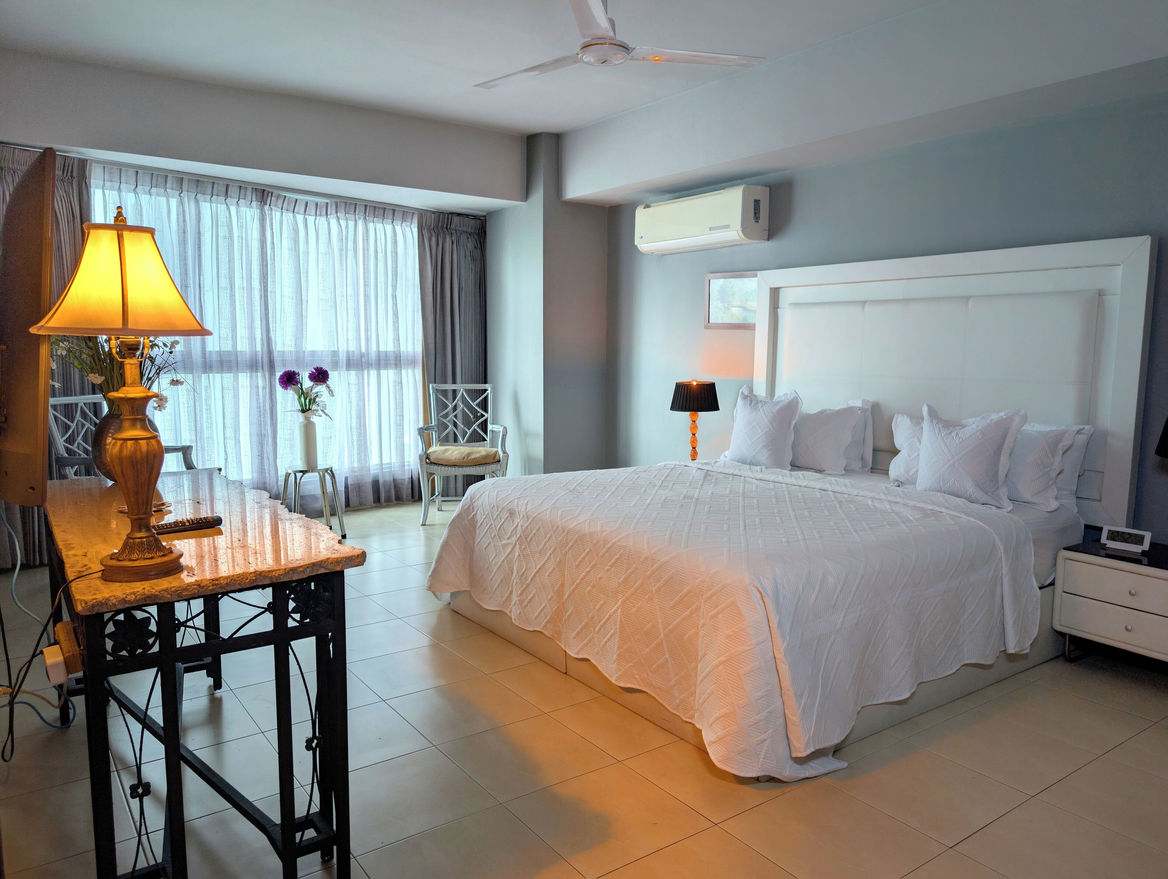 Una habitación de hotel con una cama grande, un escritorio con lámpara, una silla, un ventilador de techo y un aire acondicionado.