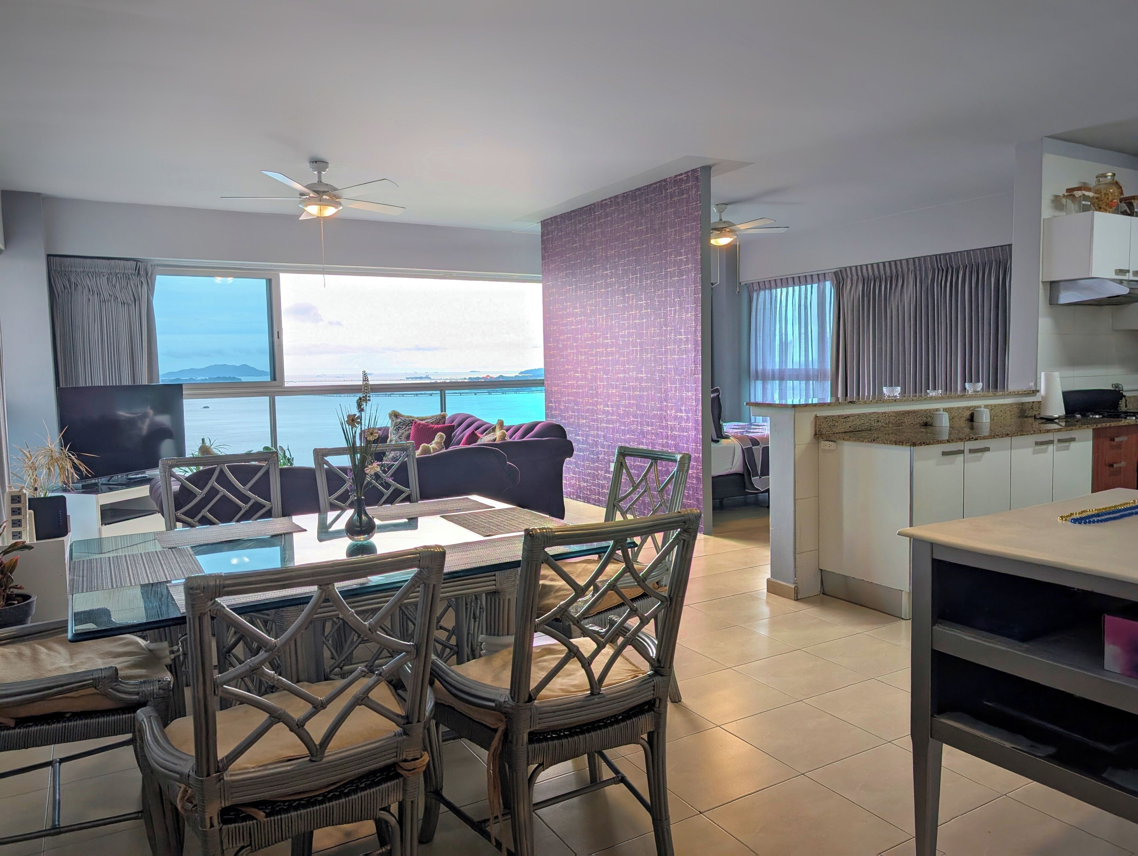 Een moderne woonkamer met een groot raam met uitzicht op zee, een eethoek met rieten stoelen, een keuken met witte kastjes en een paarse accentmuur.