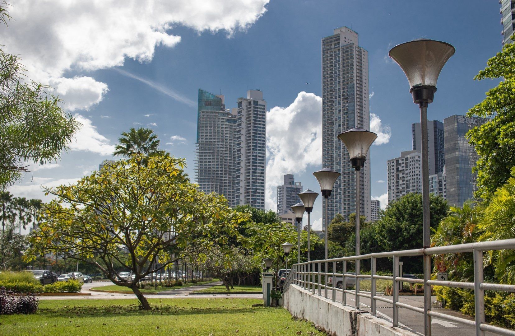 Un parque urbano con un sendero, farolas y edificios altos al fondo.