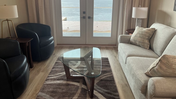 Un salón con una mesa de centro de vidrio, un sofá y una silla. Se ve una masa de agua a través de una puerta de cristal.
