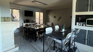 Ein Essbereich mit einem Tisch, gedeckt für vier Personen, umgeben von Stühlen. An der Wand ist ein Fernseher angebracht, und es gibt ein Fenster mit Jalousien.