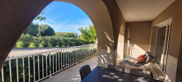 A balcony with a white railing, a table with chairs, and a view of a road and greenery.