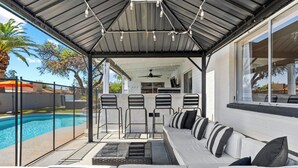 Een terras buiten met een zwembad, een zithoek en een overdeeld gedeelte met stoelen en een tafel.