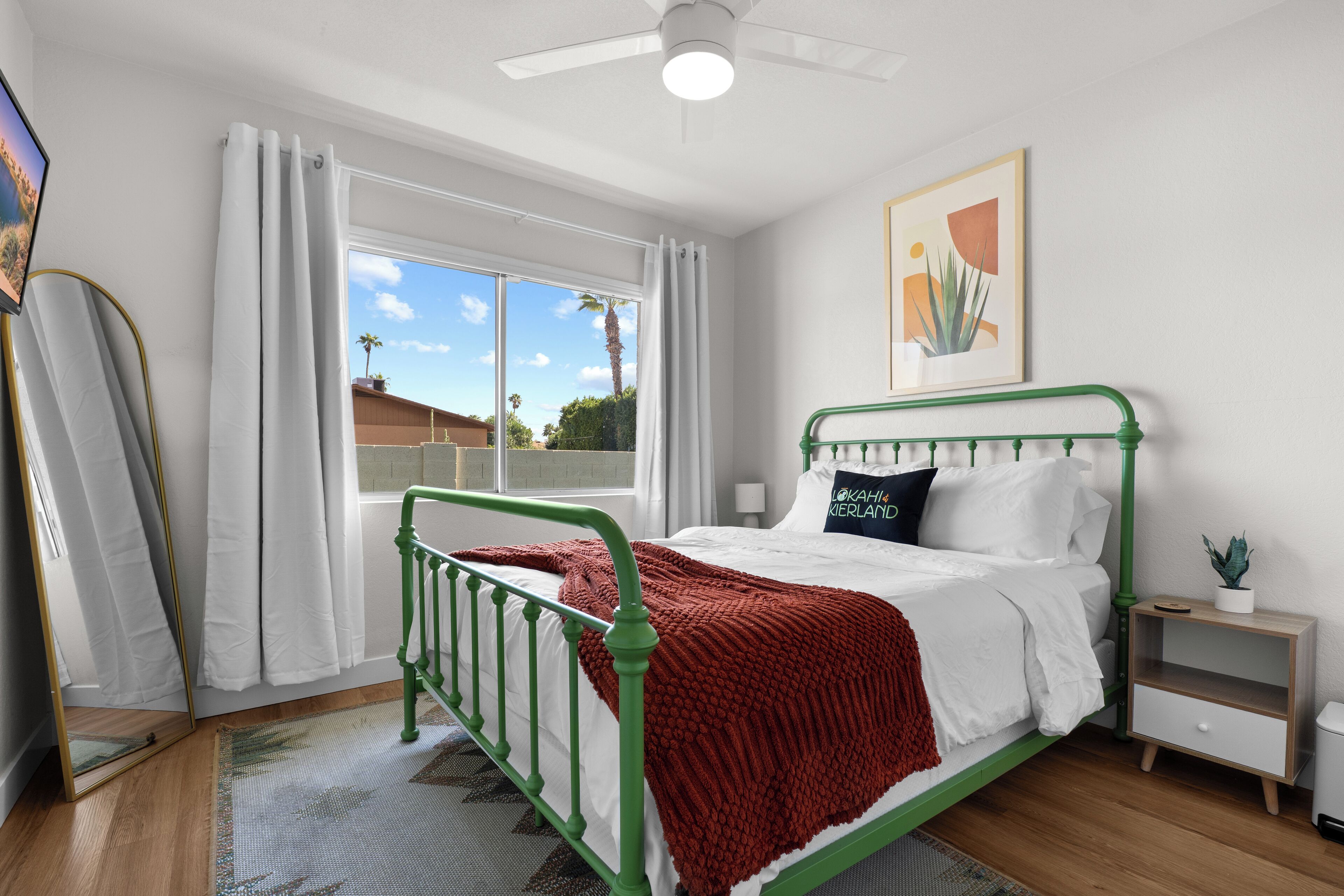 A bedroom with a green metal bed frame, white bedding, a red throw blanket, a small bedside table with a plant, and a window with white curtains.