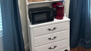 A white dresser with drawers and shelves, holding a microwave, a red coffee maker, and wicker baskets. - Historic Downtown Studio (Pensacola)