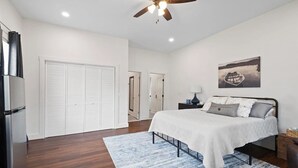 A bedroom with a bed, a ceiling fan, a nightstand, a lamp, and a painting on the wall. - Historic Downtown Studio (Pensacola)
