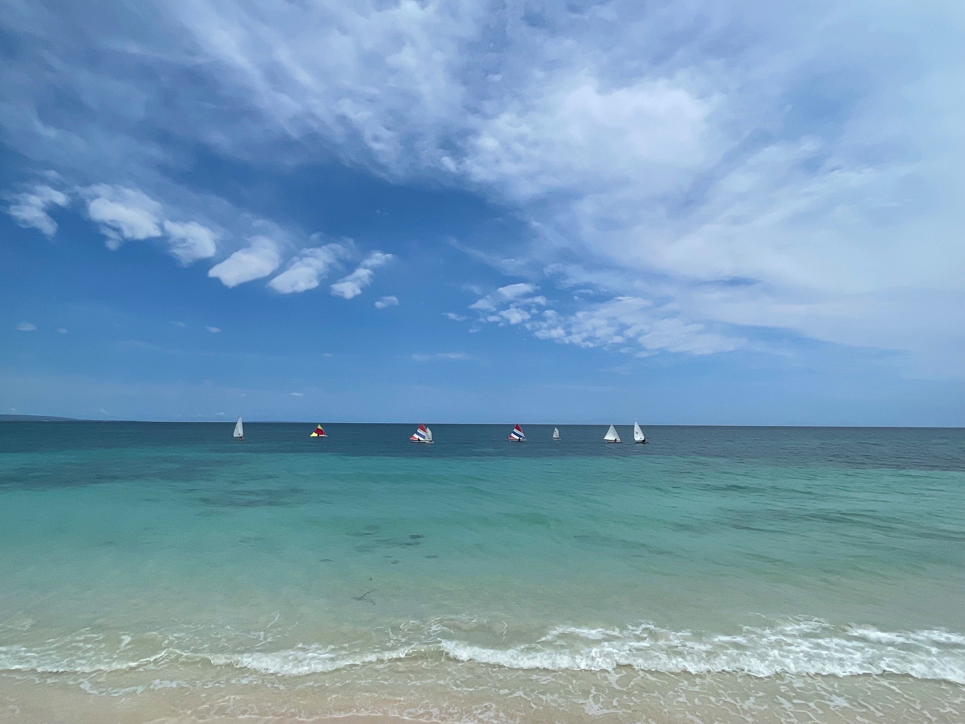A beach with clear turquoise waters and a row of sailboats.
