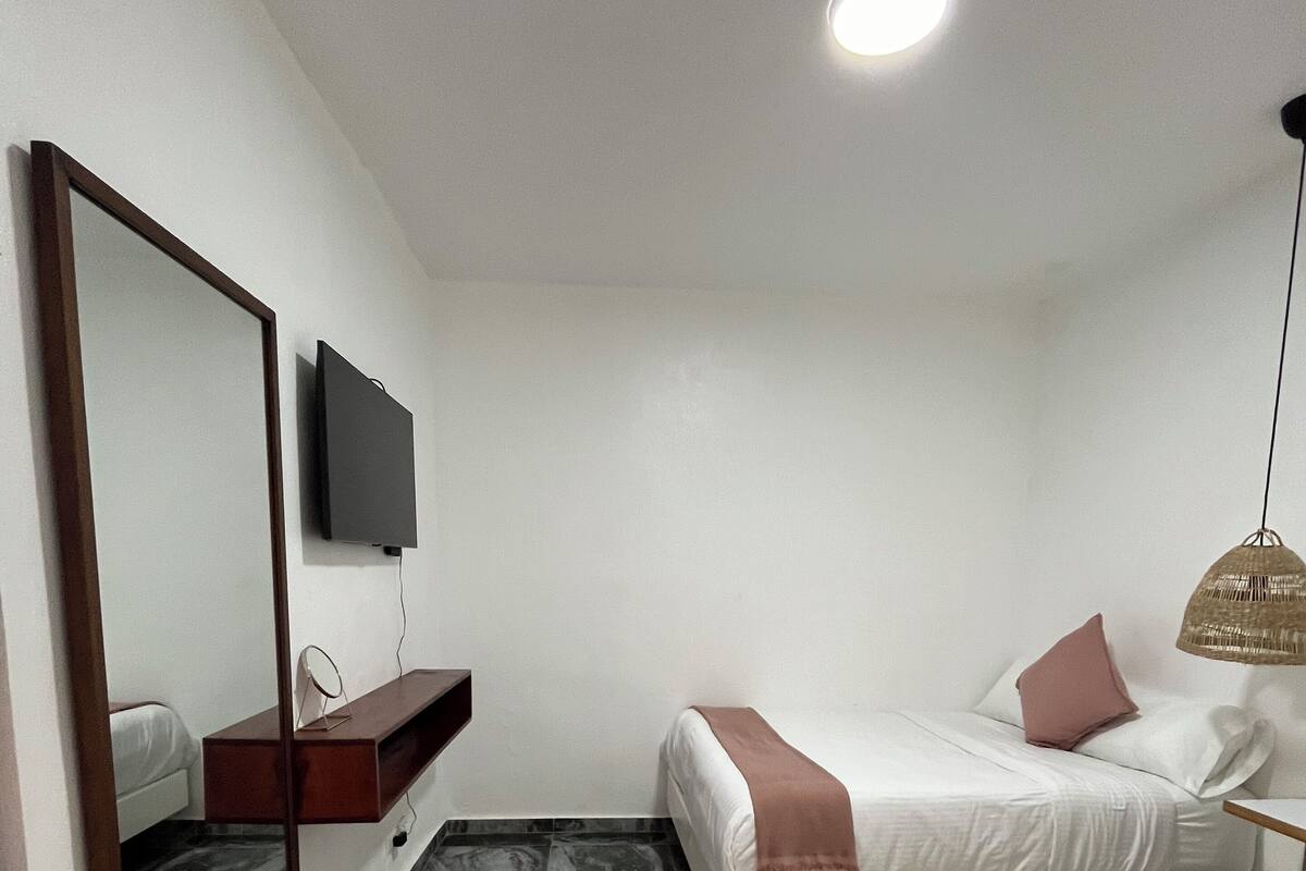 Un dormitorio moderno con un espejo grande, un televisor de pantalla plana, una mesita de noche, una cama con una manta rosa y una lámpara colgante.