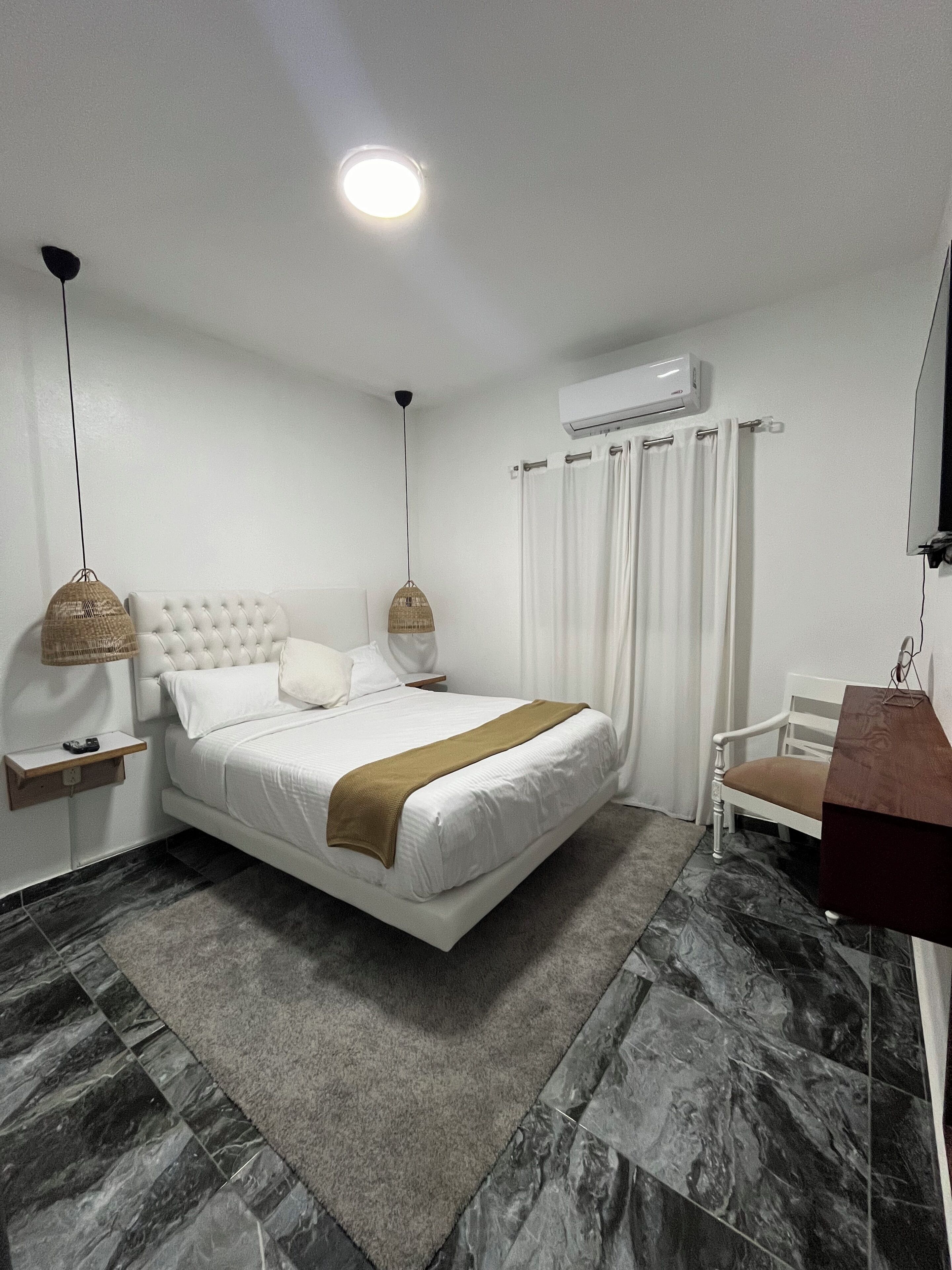 A modern bedroom with a large bed, a bedside table, a hanging lamp, a chair, and an air conditioning unit.