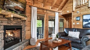 A cozy cabin interior with a stone fireplace, wooden beams, a leather sofa, and a large window overlooking a deck.