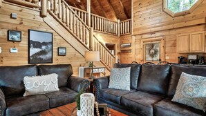 A cozy log cabin interior with a leather sofa set, a wooden coffee table, and a staircase leading to an upper level.