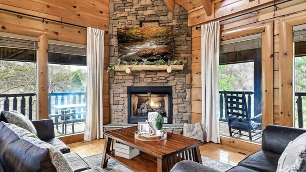 A cozy room with a stone fireplace, wooden furniture, and large windows overlooking a deck.