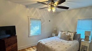 Une chambre à coucher avec un lit, un plancher en bois, un ventilateur de plafond et un téléviseur posé sur une commode.