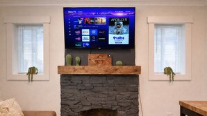 A living room with a stone fireplace, a flat-screen TV mounted above it, and two windows with blinds.