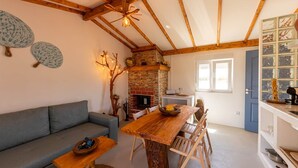 A cozy living room with a stone fireplace, wooden beams, and a dining area with a wooden table and chairs. - Monte dos Mestres (São Domingos)