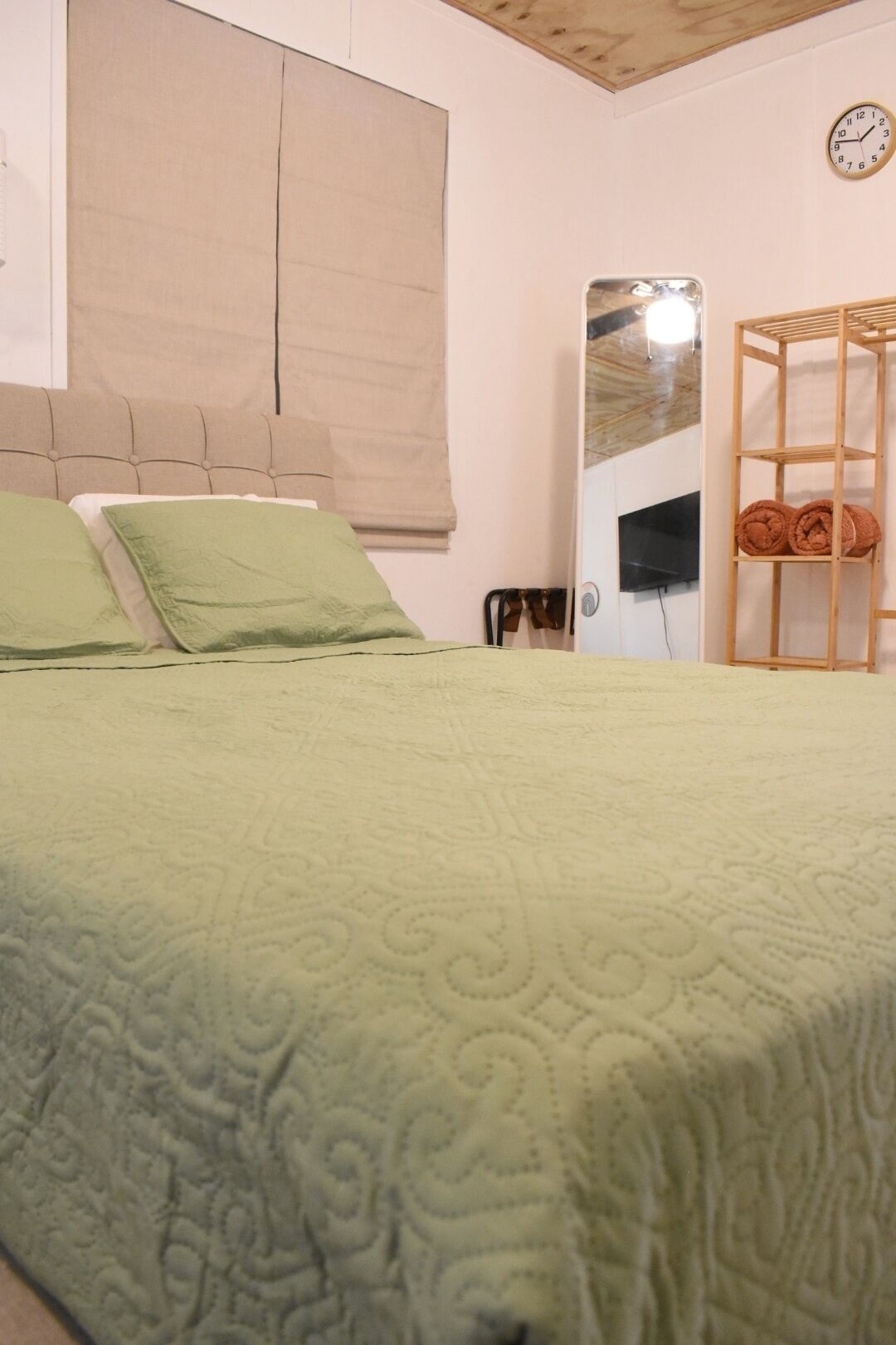 A bedroom with a bed, a wooden headboard, a mirror, a wooden shelf with towels, and a clock on the wall.