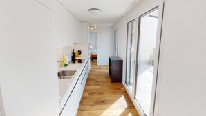 A narrow hallway with a kitchen area on one side, wooden flooring, and a sliding glass door leading to a balcony.
