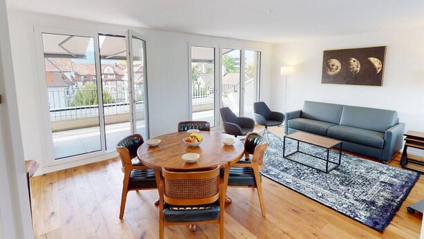 A modern living room with a round wooden dining table, two sofas, and a large window with a view of buildings.