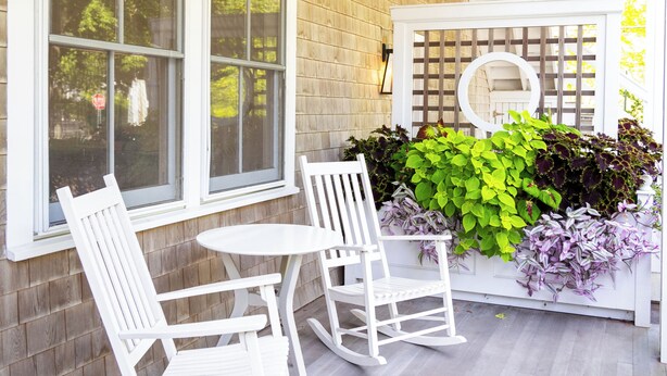 Une véranda avec des fauteuils à bascule blancs, une petite table et un jardinier contenant des plantes vertes et violettes.